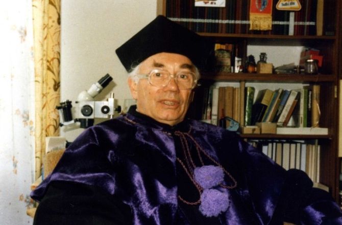 Prof. Zenon Czerniakowski siedzi ubrany we filetową togę, na głowie czarny czepek, w tle mikroskop i ksiązki