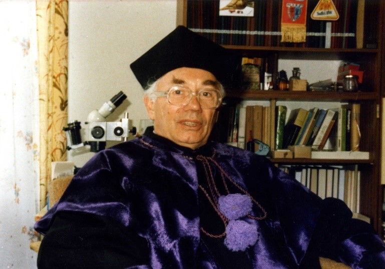 Prof. Zenon Czerniakowski siedzi ubrany we filetową togę, na głowie czarny czepek, w tle mikroskop i ksiązki